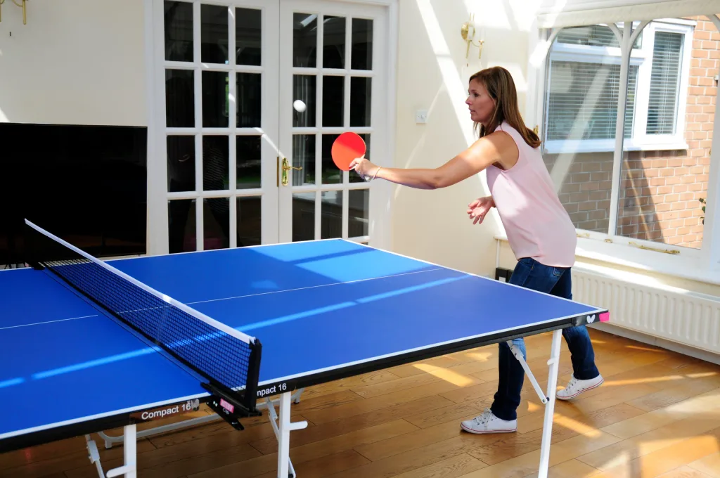 Woman in a pink top hitting a ball at a blue table tennis table with light silver legs and Compact 16 printed on the apron.