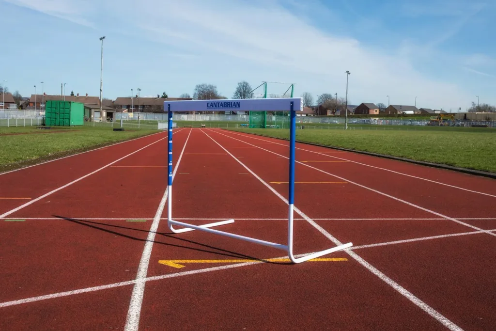 Blue and white Cantabrian training hurdle with UPVC top board standing on a red running track.