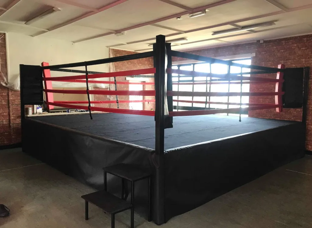 Competition ring with black canvas and skirt, red ropes and powder-coated steel corner posts in a brick-walled training space.
