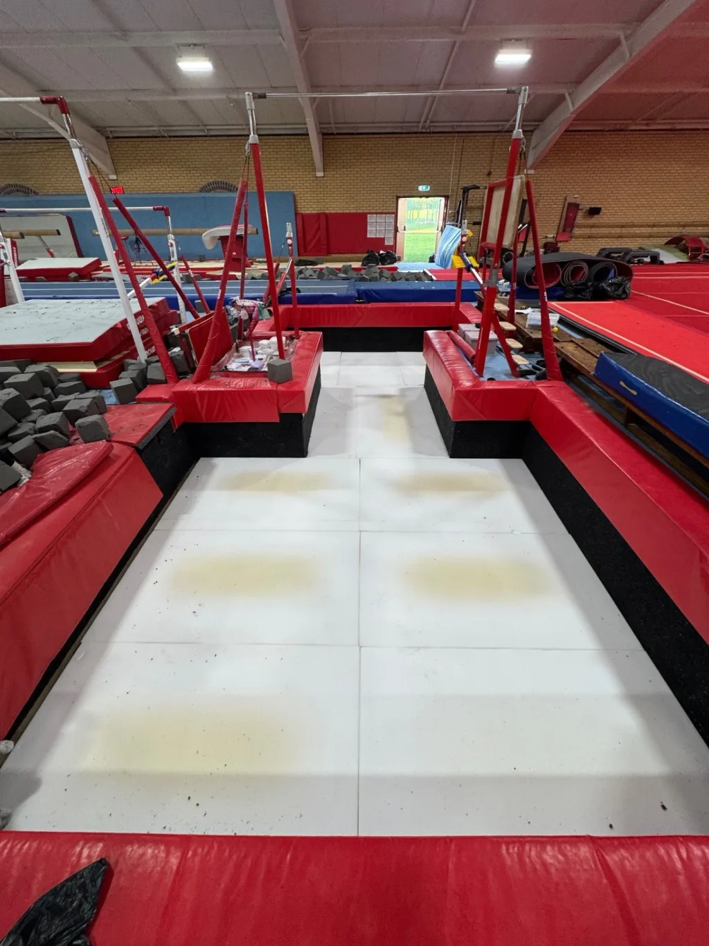 White gym pit base foam panels with pale yellow stains installed at the bottom of a foam pit, surrounded by red padded edging and gymnastics bars above.