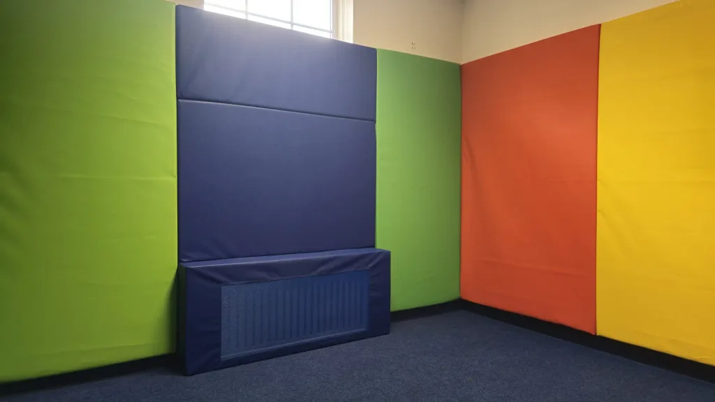 Corner of a padded room with green, blue, orange and yellow wall pads and a blue padded radiator cover on a blue carpet beneath a high window.