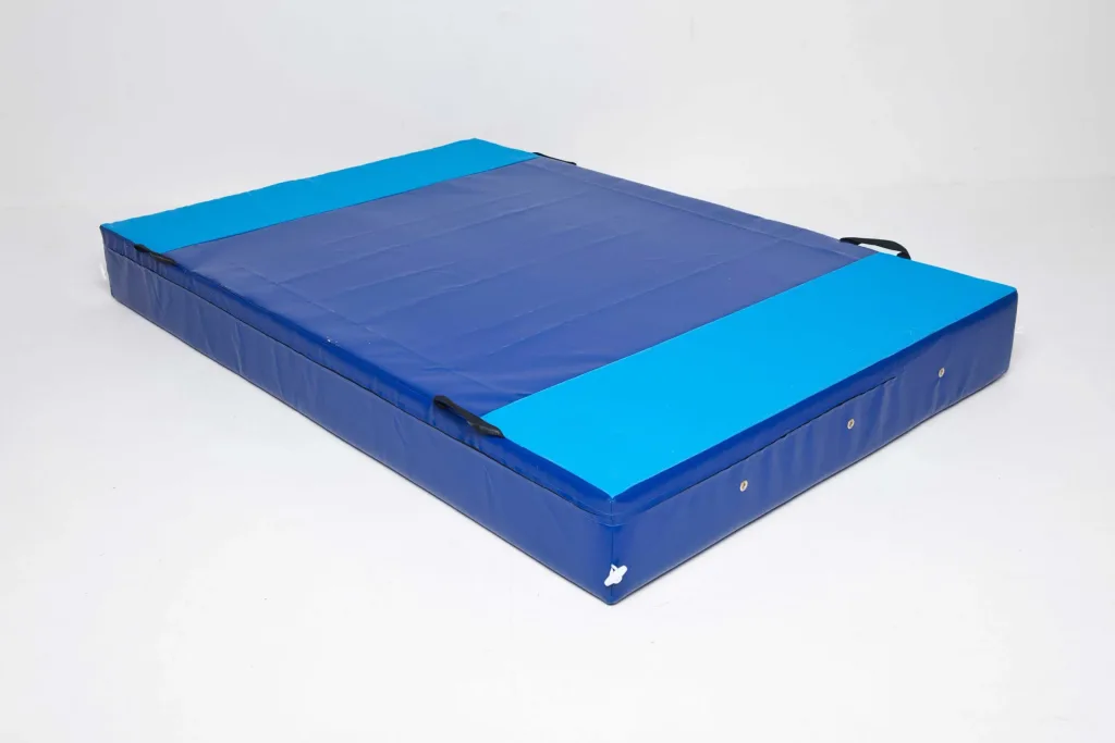 Blue crash mat with light-blue end panels, dark-blue top, black carrying handles and metal grommets on a white studio floor.