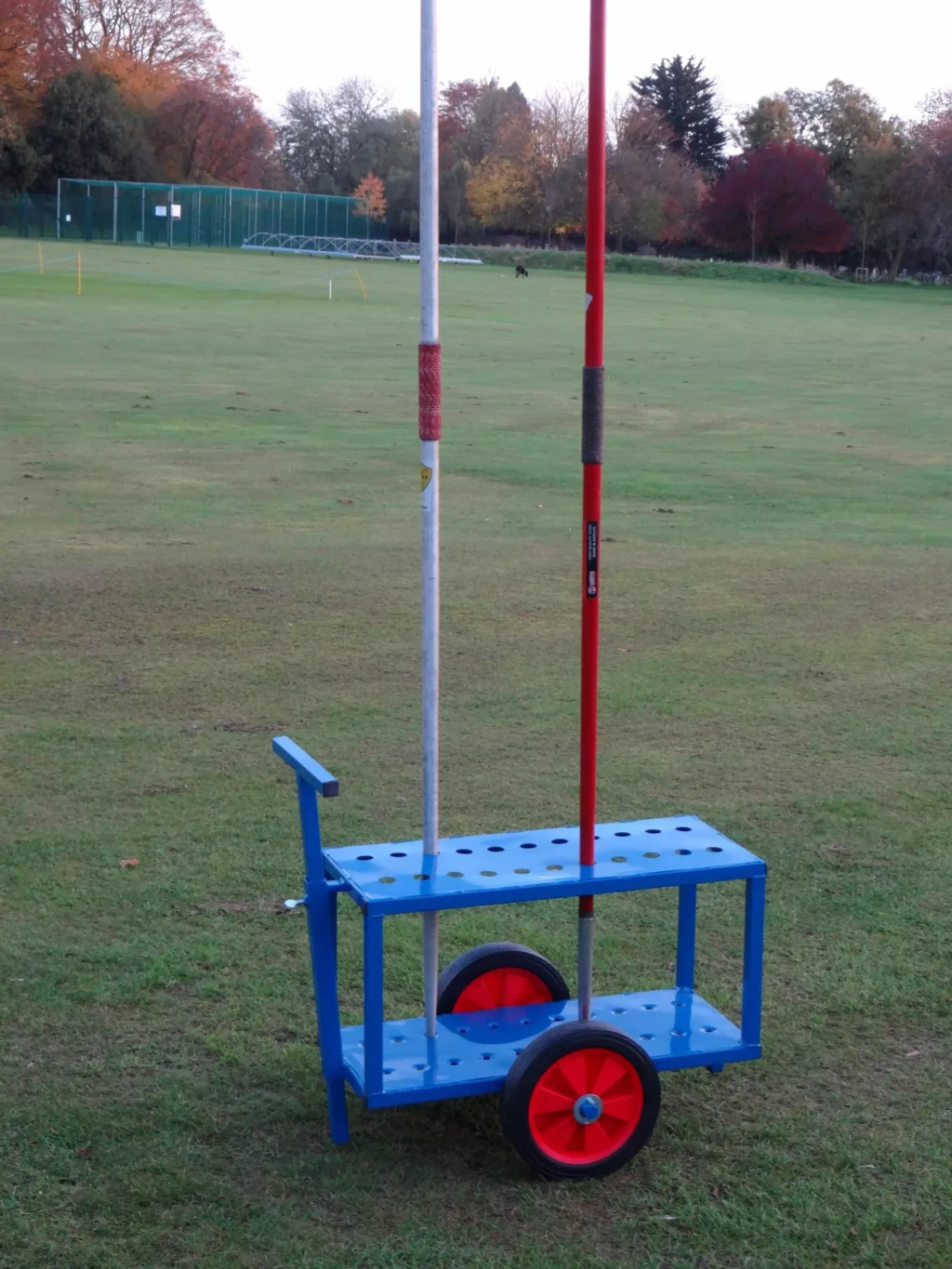 Blue powder-coated steel javelin trolley with two upright javelins and large hard rubber red wheels on a grassy field.