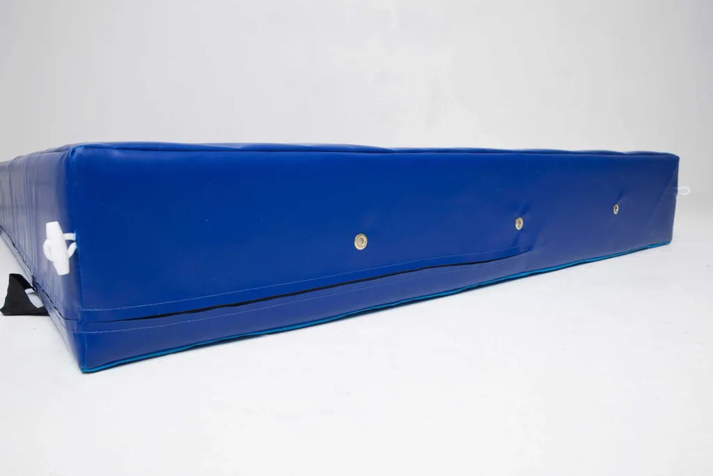 Blue crash mat with a zipped nylon-reinforced PVC cover, brass grommet breather holes, anti-slip base panels and a visible white rope handle.