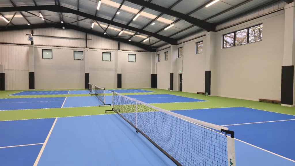 Indoor sports hall with multiple light blue courts and green surrounds, nets across the foreground, and black square post pads on the wall columns.