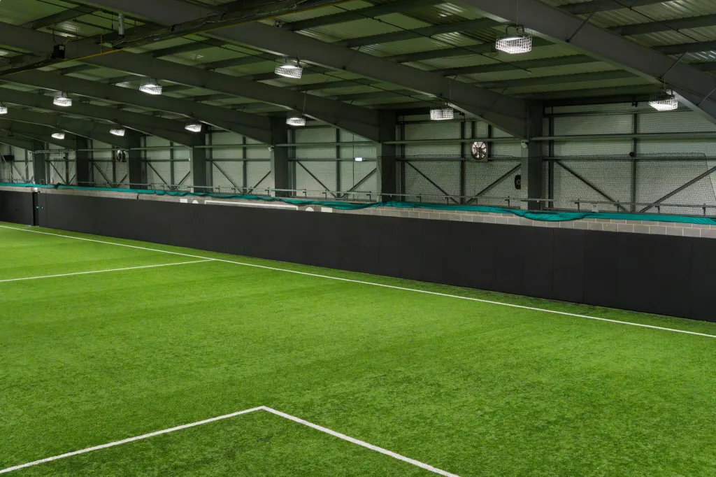 PVC-covered sports pitch safety padding panels lining an indoor artificial turf pitch wall beneath steel roof trusses and overhead lights.