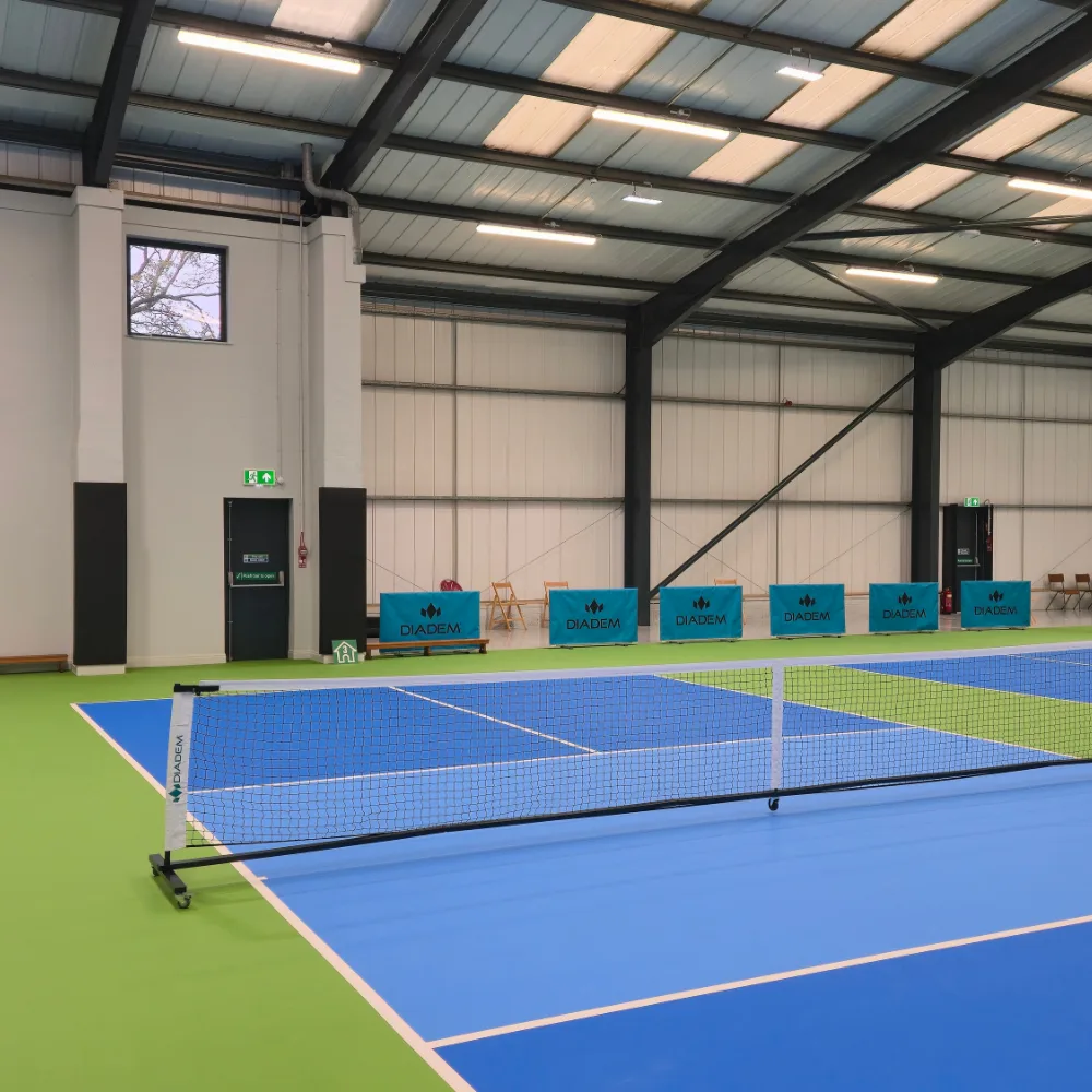 Indoor pickleball court with blue playing surface and green surrounds, net in foreground and padded wall panels with teal advertising banners along the far wall.