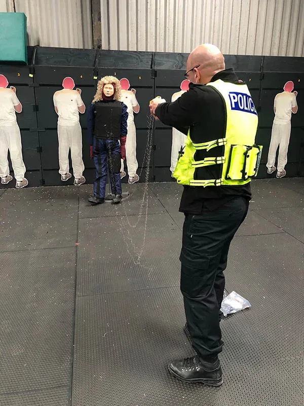 Police officer in a high-visibility vest labeled POLICE aims a training device at a manikin wearing a protective vest and leg guards.