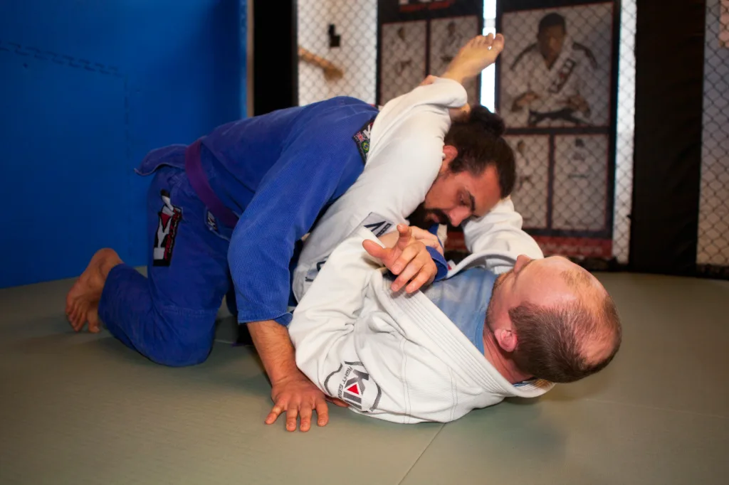Two judo practitioners in a blue gi and a white gi grappling on a tatami PVC mat beside a chain-link training cage.