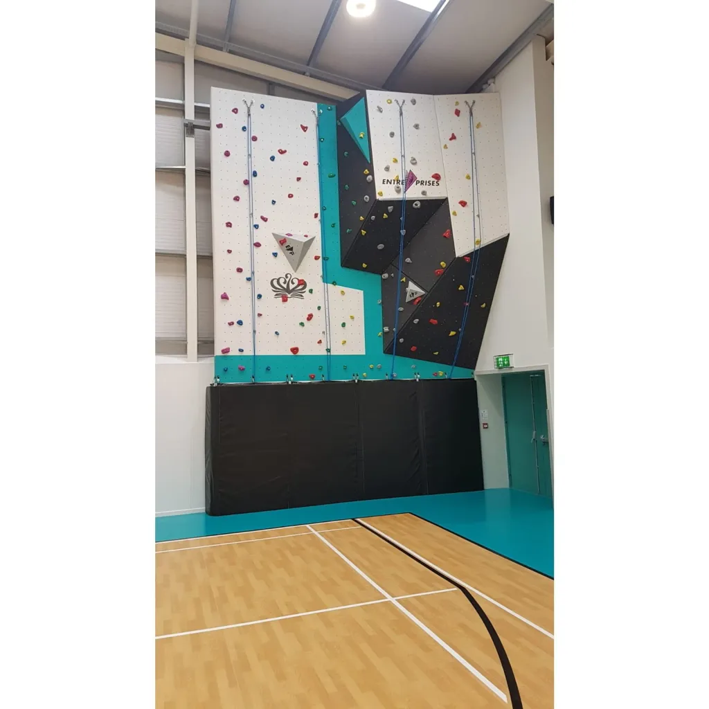 Climbing wall with teal and white panels, multicolored holds, folded black articulated landing mats at the base, and the text ENTREPRISES on a panel.