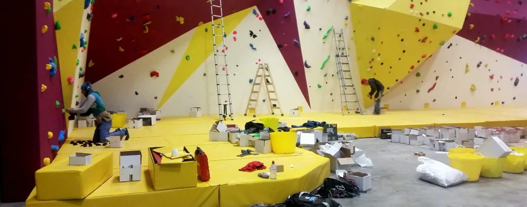 Two workers fitting colorful climbing holds on angled purple, yellow and white walls above yellow crash mats with ladders and boxes.