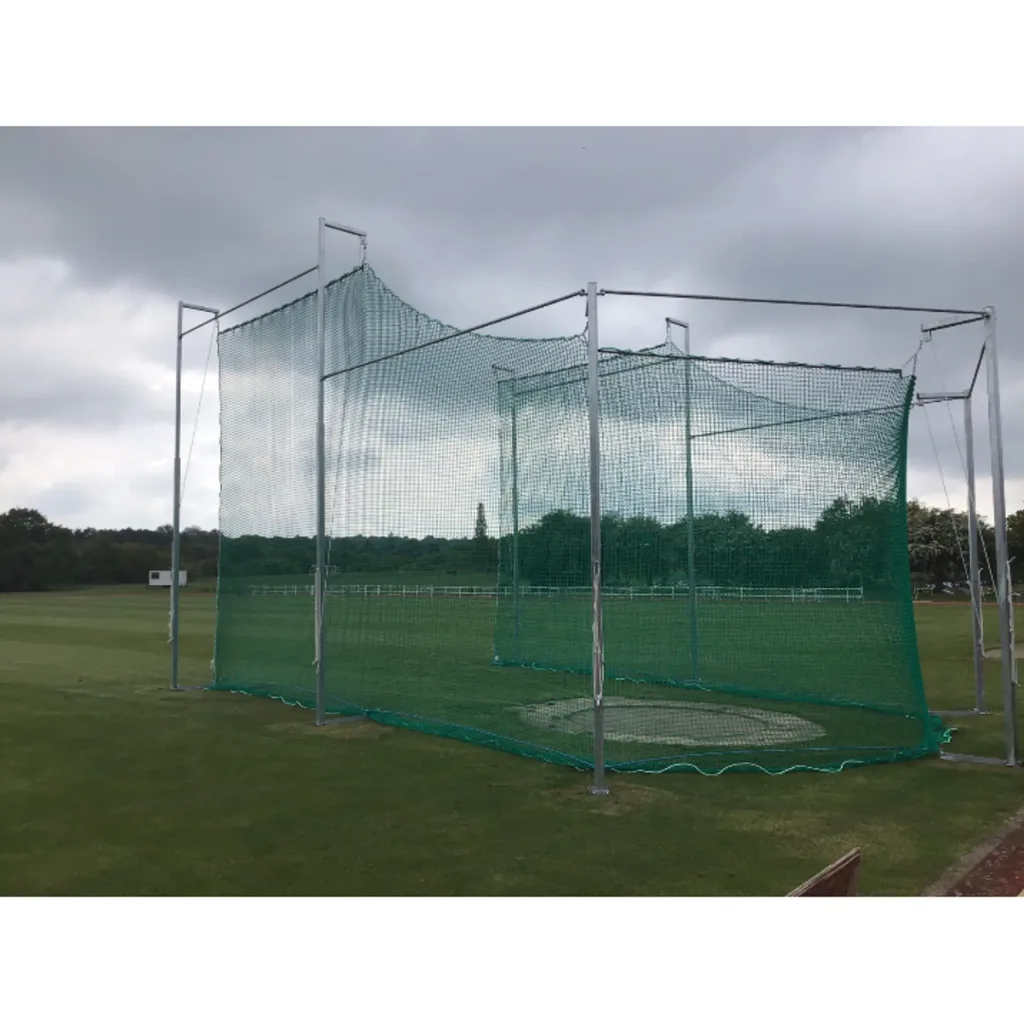 Discus cage with seven galvanised steel poles and knotless green mesh net enclosing a throwing circle on grass under cloudy sky.