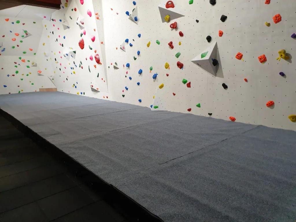 Grey carpeted crash matting with a thick foam base and no gaps under a white bouldering wall with multicolored holds.