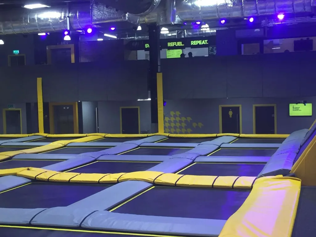Yellow heavy-duty PVC padding over foam surrounds rows of blue trampoline beds in an indoor park, with a lit sign reading REFUEL. REPEAT.