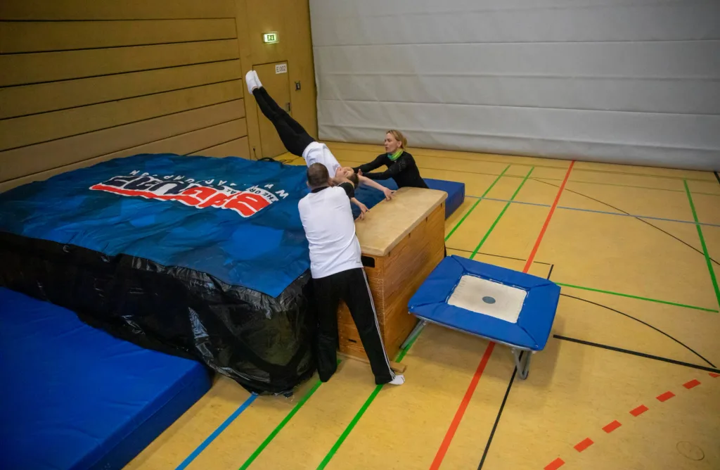 Person mid-flip into a large blue inflatable mat while two instructors steady them beside a wooden vault, door sign reading E 002.