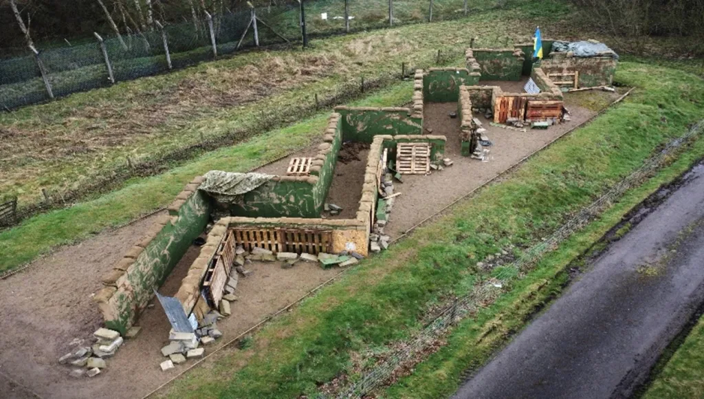 Aerial view of an outdoor tactical training layout with green modular walls topped with sandbags, wooden pallets, and gravel pathways.