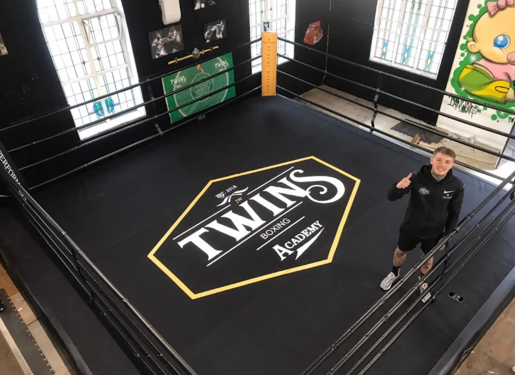 Raised platform training ring with black canvas featuring the text TWINS BOXING ACADEMY inside a yellow hexagon and a man giving a thumbs-up.