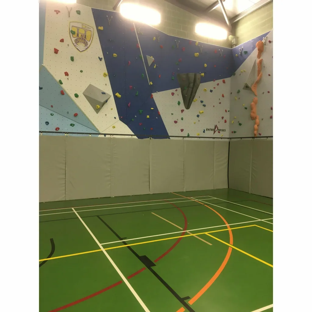 Gray articulated climbing mats folded upright along the base of an indoor climbing wall above a green sports hall floor.