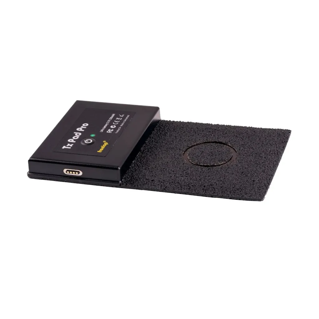 Black Tx Pad Pro transmitter resting on a black textured rectangular mat with a circular contact area and a side charging port.