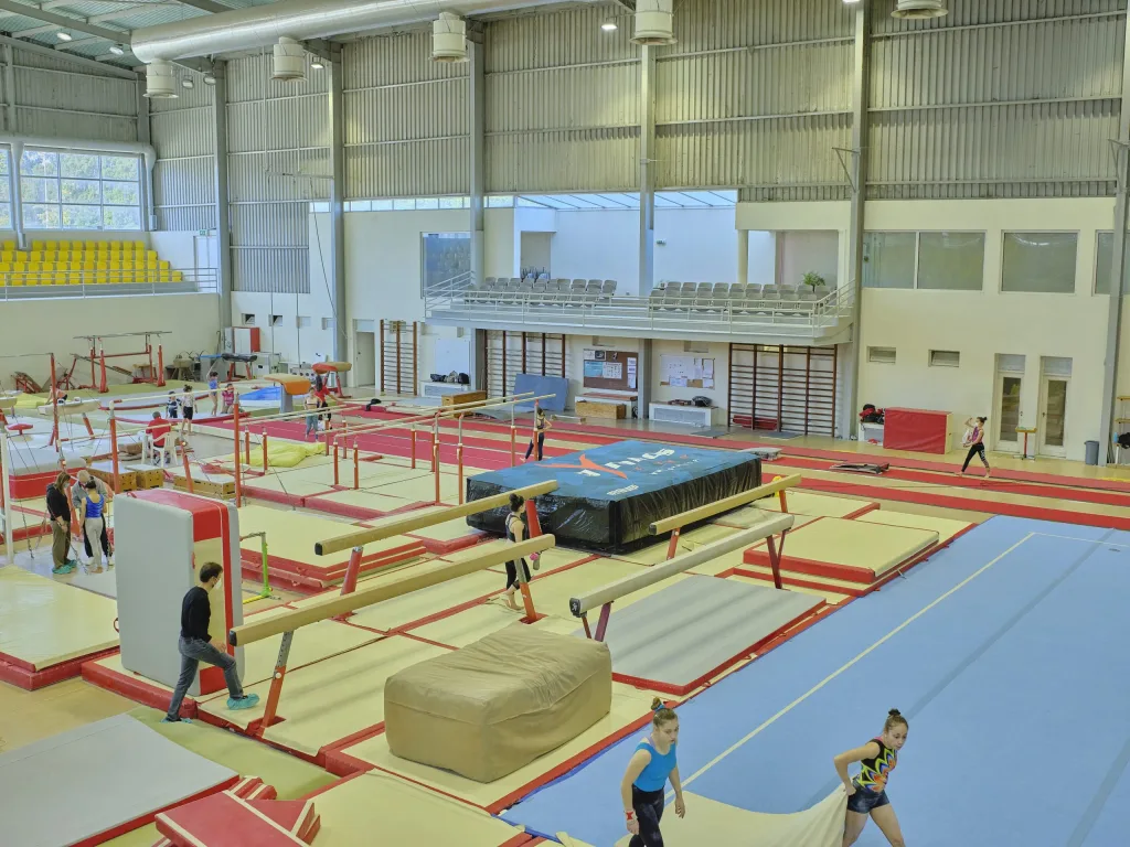 Indoor gymnastics hall with blue tumbling floor, multiple balance beams, a large black covered landing bag and athletes practicing.