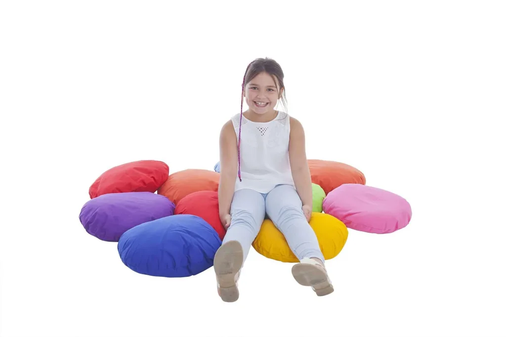 Girl sitting on ten cotton scatter cushions arranged in a flower shape in random bright colours, smiling.