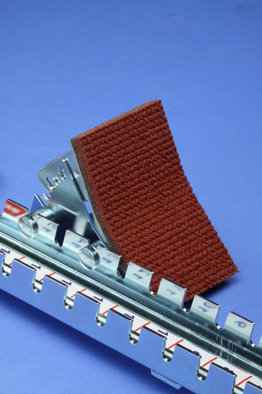 Competition galvanised steel starting block with a red textured footplate pad mounted on a notched centre bar against a blue background.