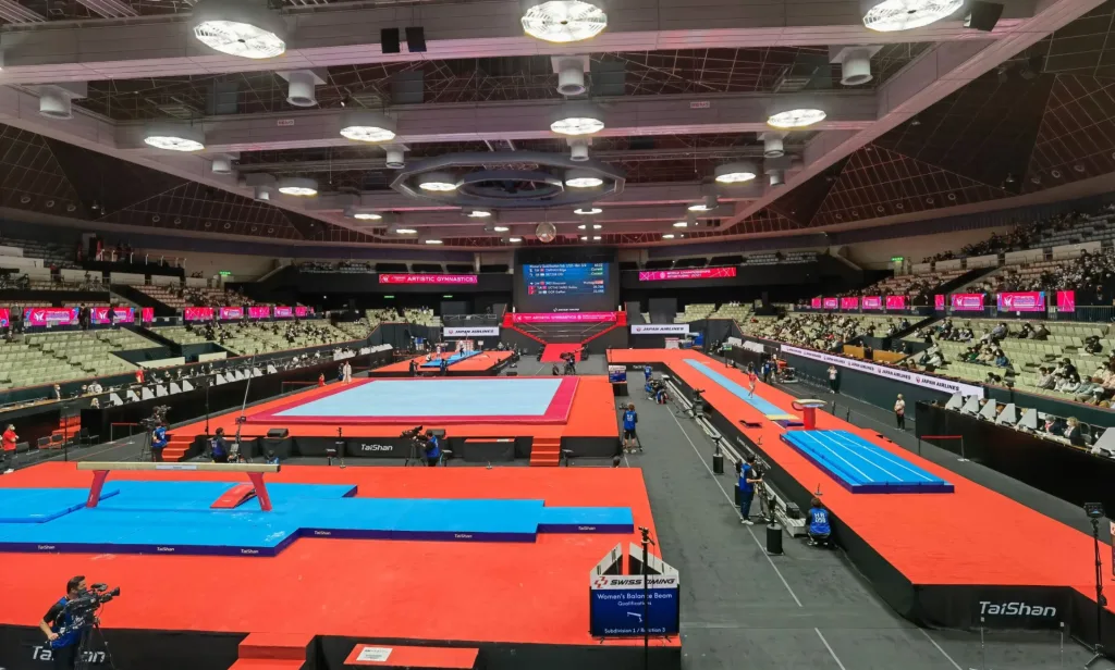 Large indoor gymnastics arena with red competition floor, blue mats and a balance beam, a central scoreboard and pink banners reading Artistic Gymnastics.