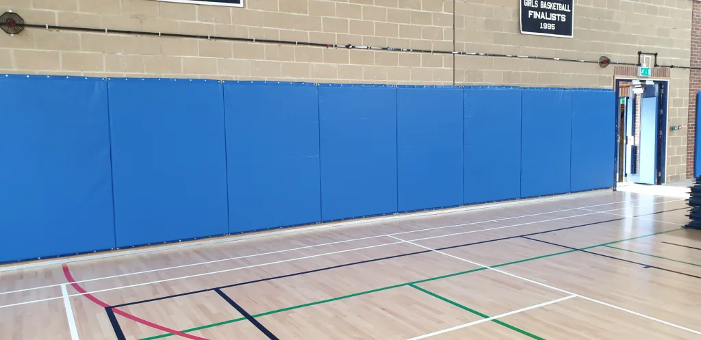 Blue wall pads line a gym wall above the marked wooden floor, with a sign reading GIRLS BASKETBALL FINALISTS 1995.