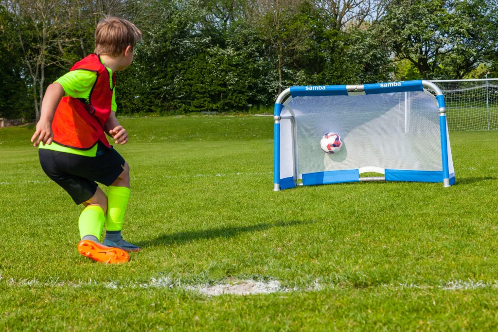 Child kicking soccer ball into small blue folding aluminium goal labeled samba on a grassy pitch.