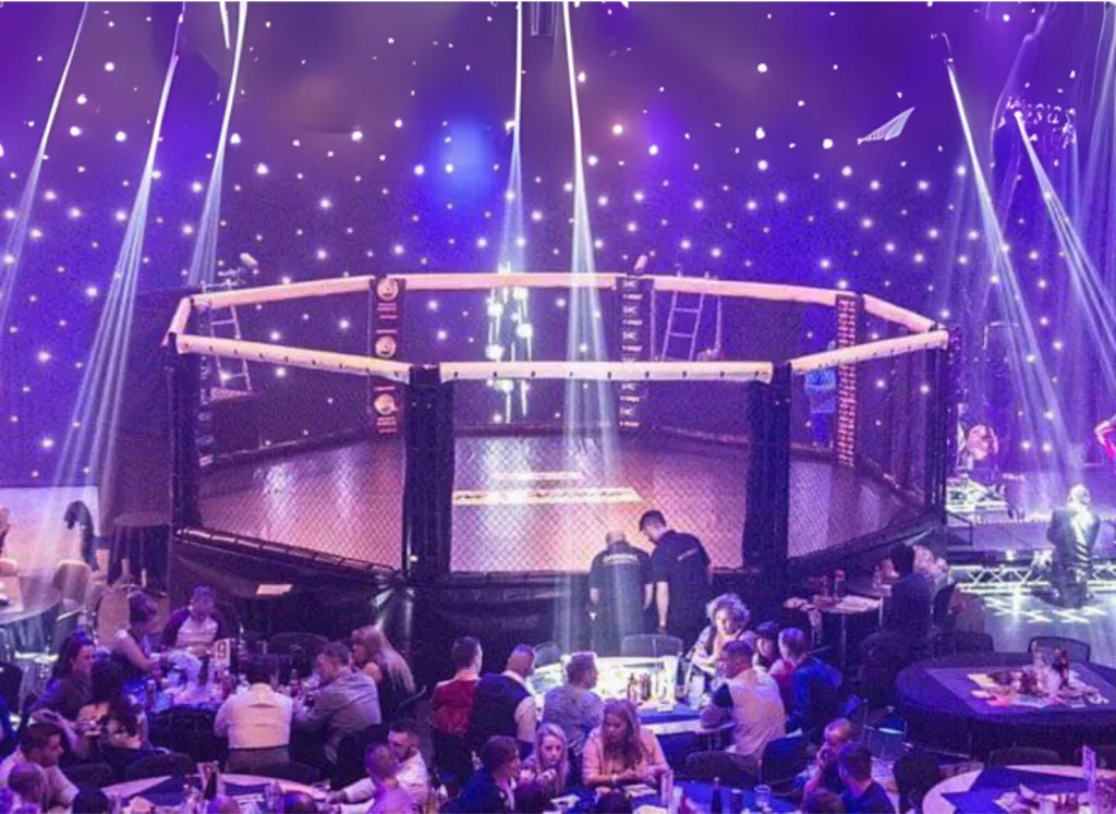 Competition cage with raised deck, steel powder-coated frame, black mesh and padded rails, illuminated by purple stage lights above seated audience.