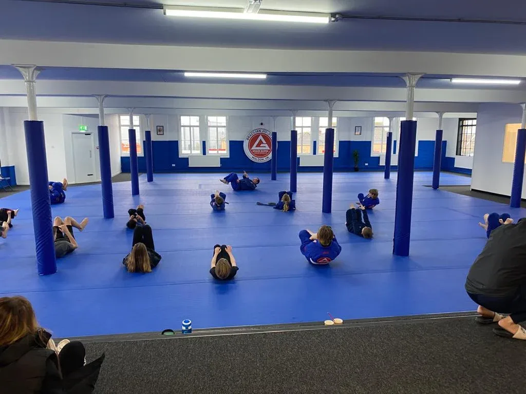 People doing floor exercises in a training hall on large blue PVC roll-out mats with padded blue columns.