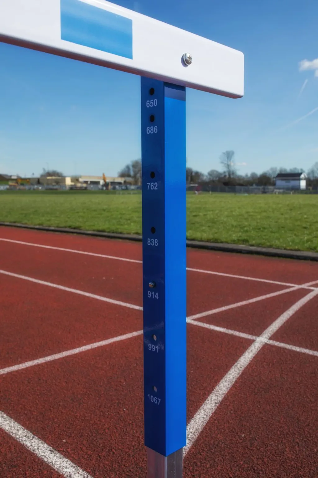 Blue steel hurdle upright with white top bar showing height markings 650, 686, 762, 838, 914, 991, 1067 on a red running track.