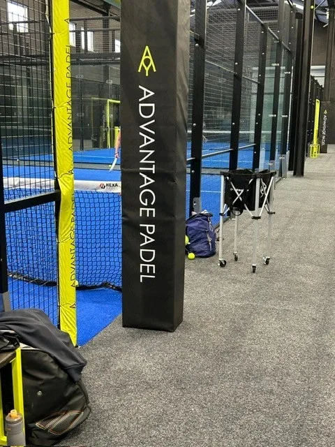 Black heavy-duty PVC post padding with white vertical text beside a yellow net post on a blue padel court, with bags and a ball cart nearby.