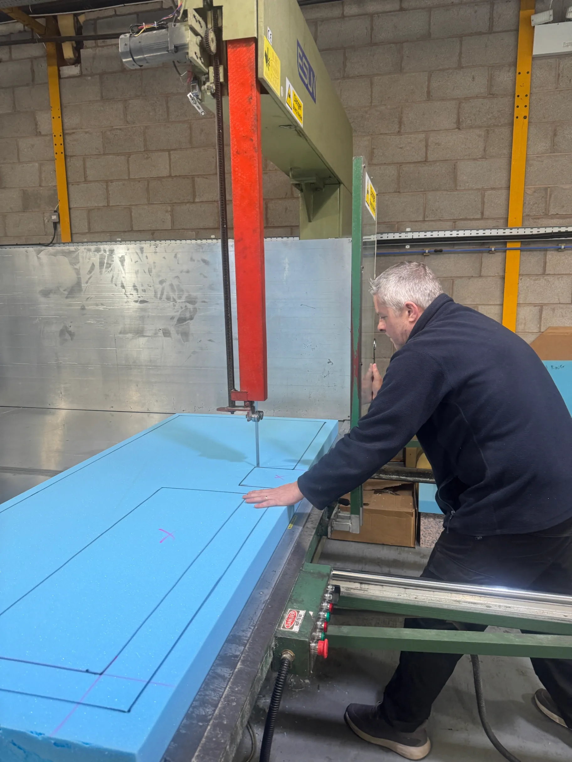 Cutting Process Man operating a vertical bandsaw cutting a large blue foam sheet along drawn lines, with a red machine column and concrete block wall in the background.