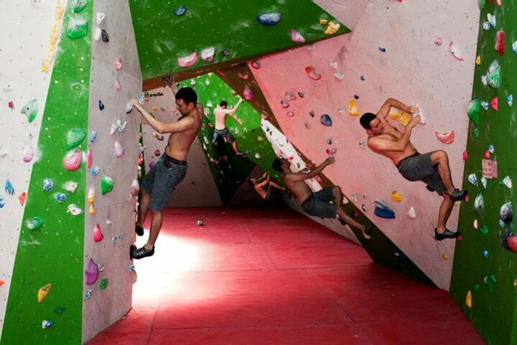 Three shirtless climbers on angled indoor bouldering walls above a thick PVC bouldering mat.