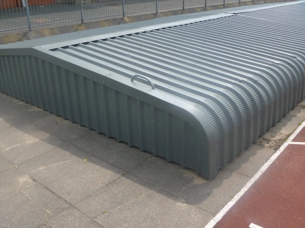 Steel wheelaway sandpit cover with corrugated epoxy painted cladding, ergonomic handle and padlock loop, positioned beside a running track.