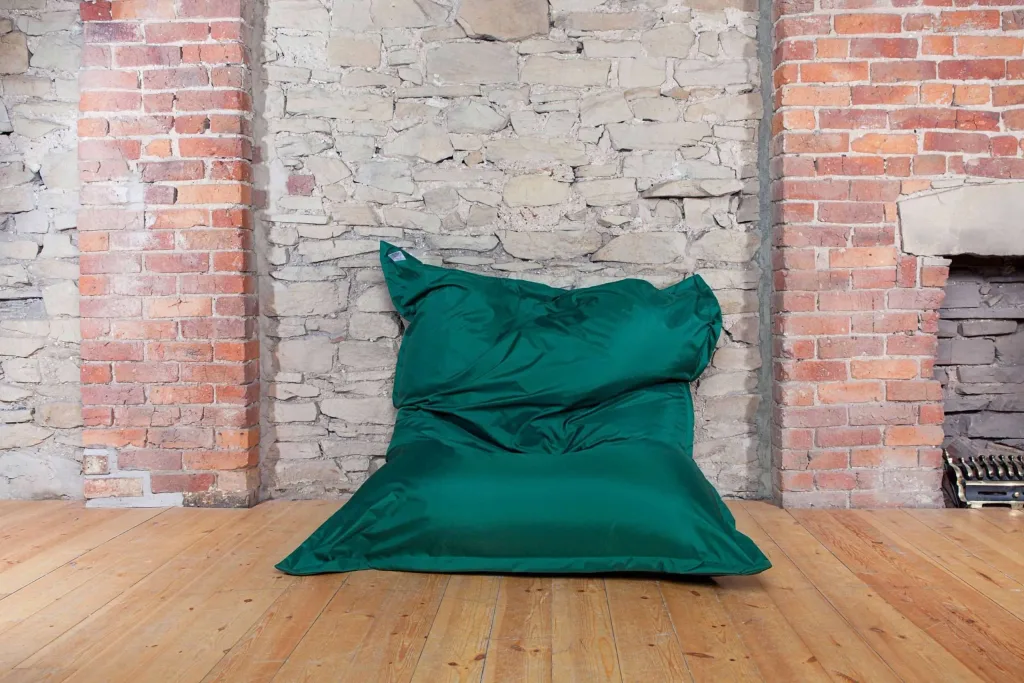 Large green polyester floor cushion for teen/adult sitting on a wooden floor against a rough stone wall with brick pillars.