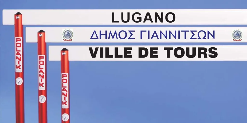 White plastic hurdle top boards labeled LUGANO, ΔΗΜΟΣ ΓΙΑΝΝΙΤΣΩΝ and VILLE DE TOURS mounted on red posts.
