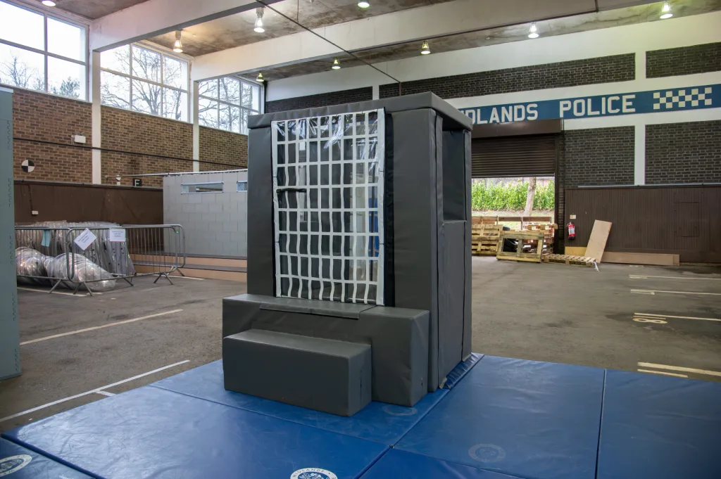 Gray reinforced-foam van cage with heavy-duty PVC cover and clear grid door on blue mats inside a training hall with 'POLICE' signage.
