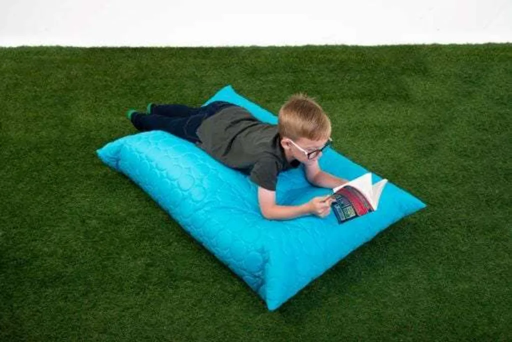 Child lying on an aqua giant child floor cushion, reading a book on green artificial grass.