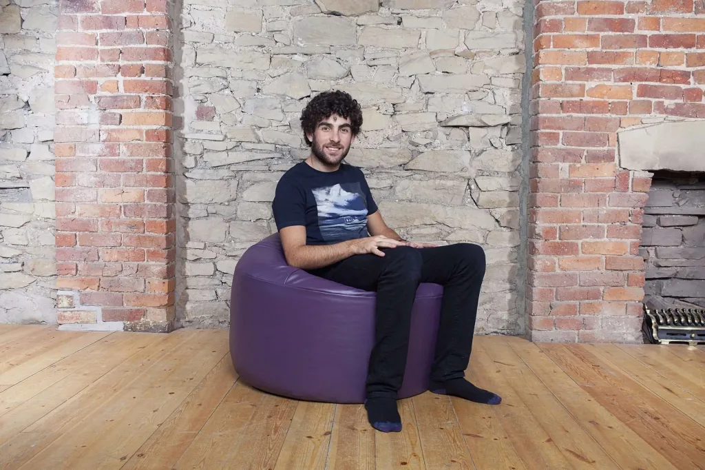 Man sitting on a purple faux leather bean bag chair in front of a brick and stone wall.