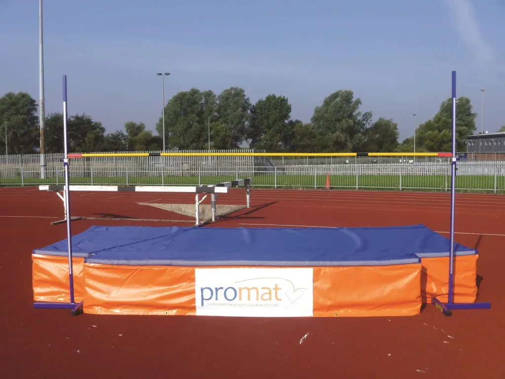 Blue-topped orange-sided high jump landing mat on a red track with purple uprights and a yellow crossbar, white label reading "promat".