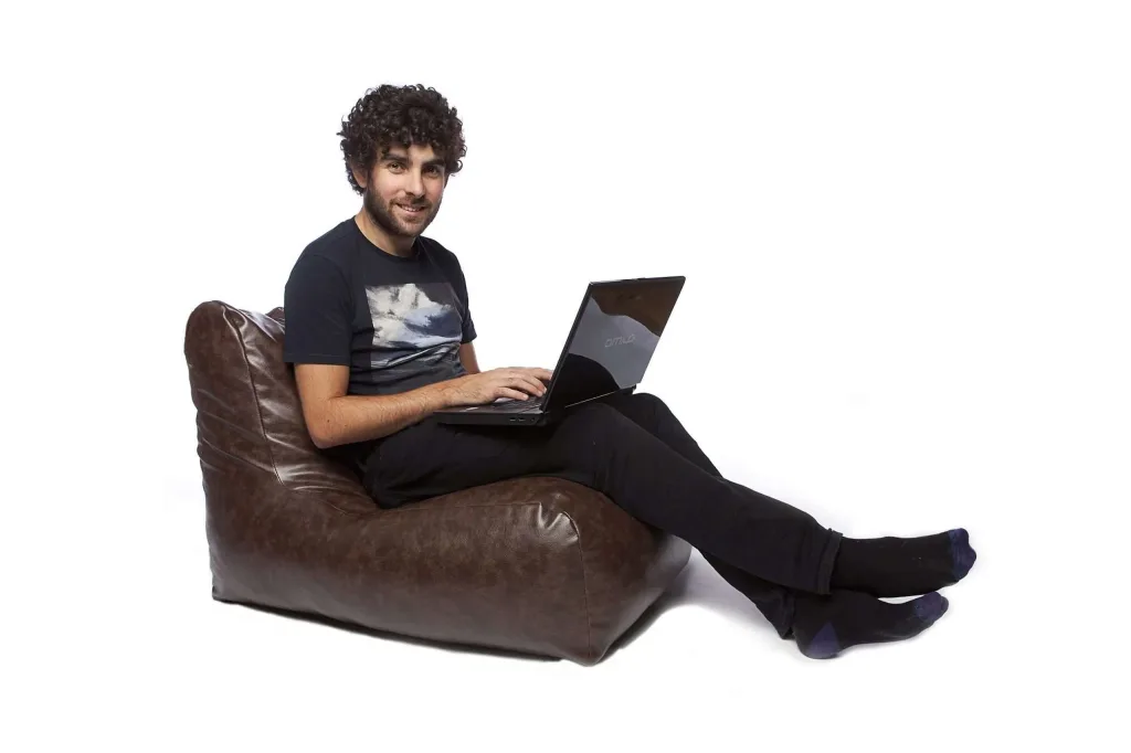 Man seated on a brown faux leather bean chair using a laptop, legs stretched out and smiling.