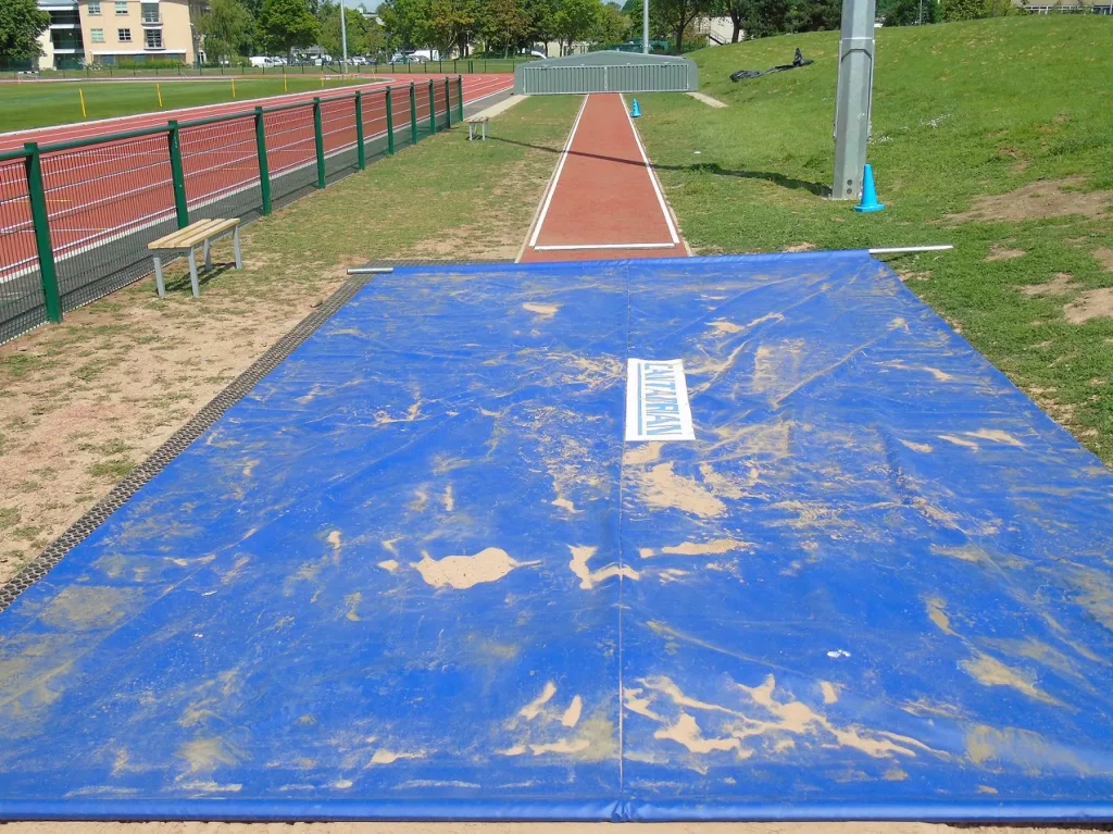 Blue PVC and mesh sandpit cover 9.3m x 3.3m spread over a long-jump pit with sandy smears and a running track beyond.