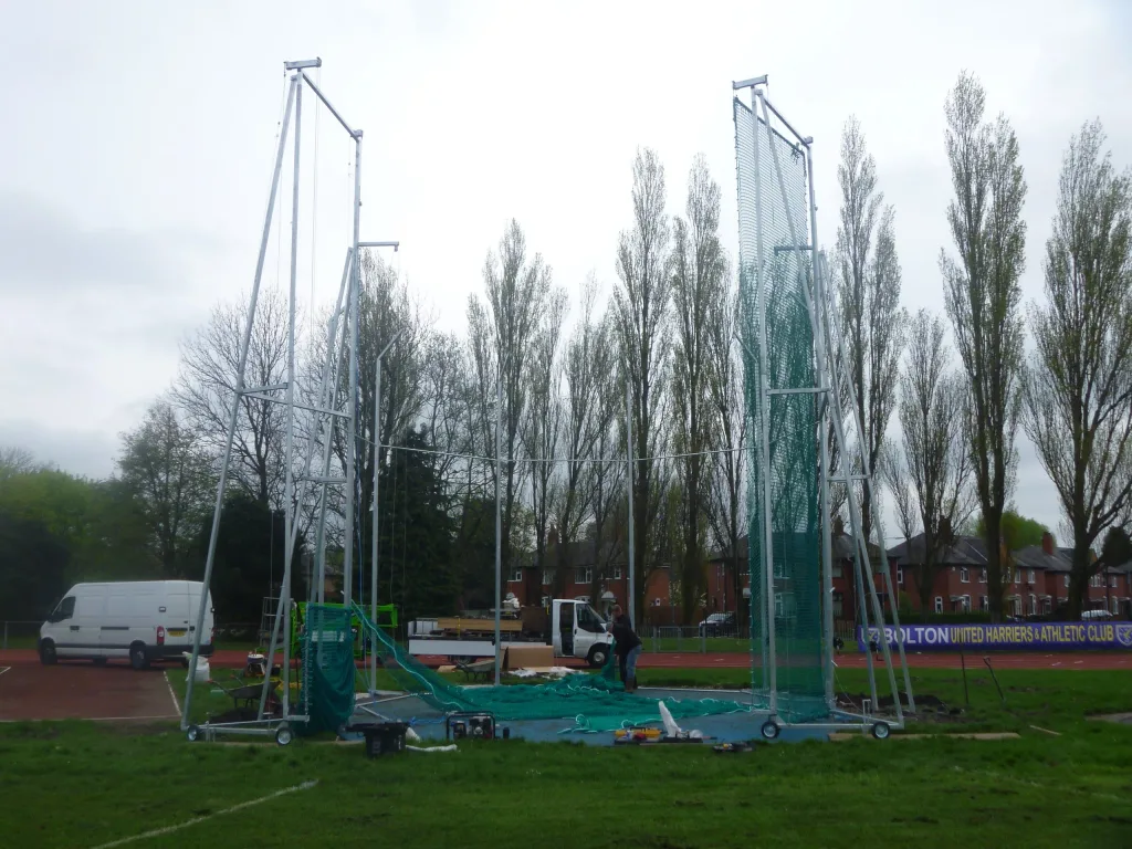Discus cage structure with green knottless netting being installed on a track field, two workers and vans nearby; banner reads BOLTON UNITED HARRIERS & ATHLETIC CLUB.