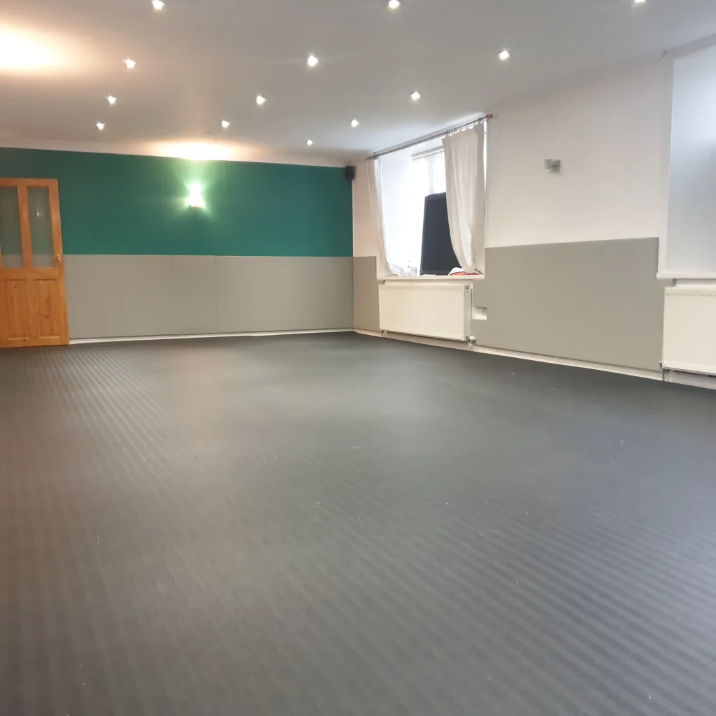 Dark gray ribbed roll-out mats cover the floor of an empty training room with light gray wall padding, a teal accent wall, wooden door, and windows.