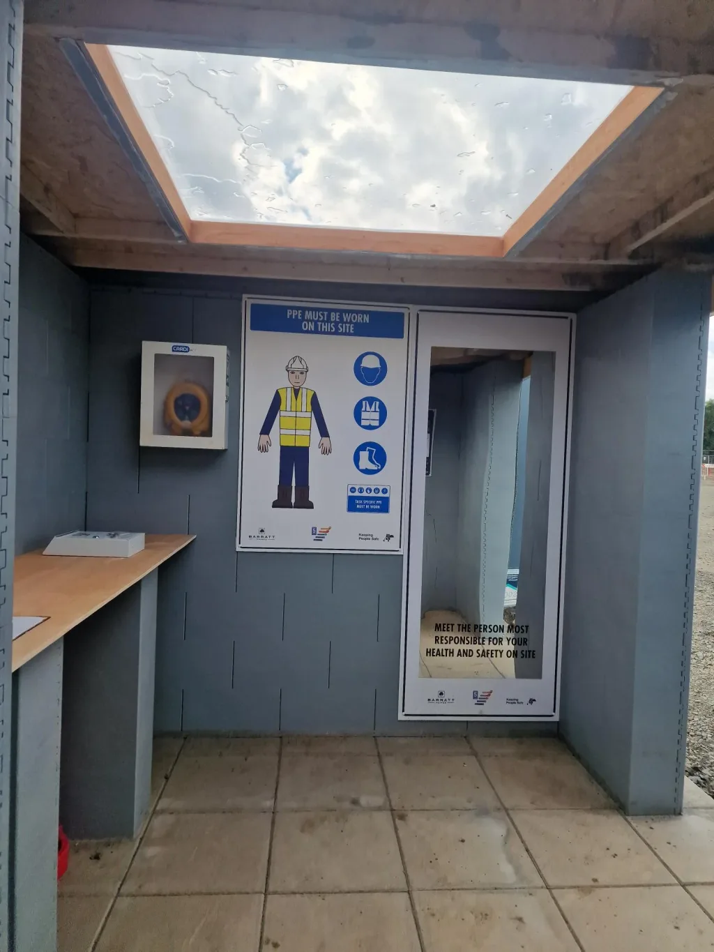 Small gray modular training room with skylight, wooden counter, and sign reading "PPE MUST BE WORN ON THIS SITE".
