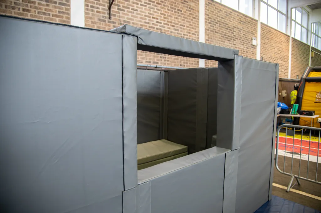 Gray padded custody cell with open viewing window and foam bench inside, made from foam wrapped in heavy duty PVCs, against a brick wall.