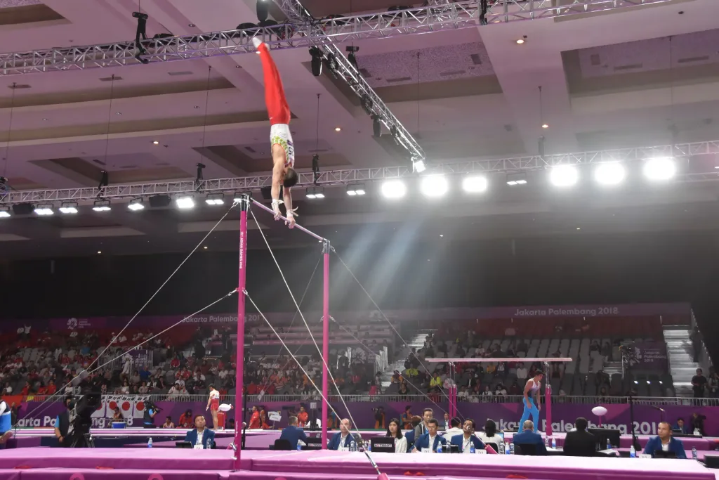 Gymnast in red pants handstanding on a high bar above pink uprights, judges' table below and banner reading 'Jakarta Palembang 2018'.