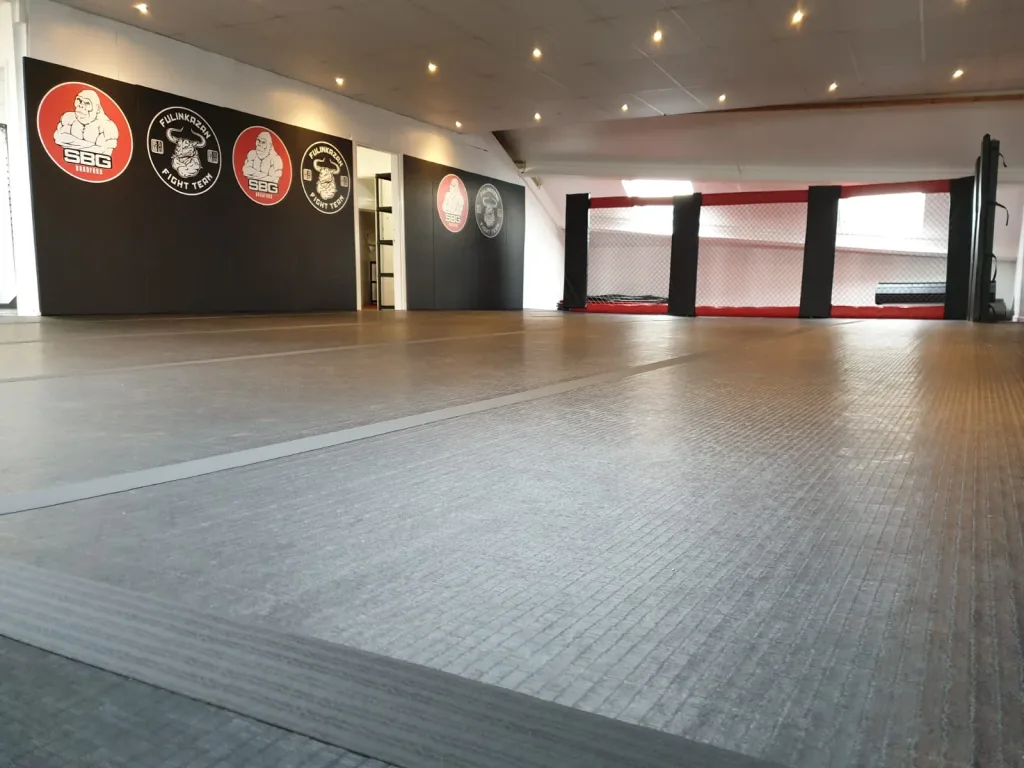 Grey hybrid roll out mats covering a martial arts training area with black wall logos and a red-and-black cage at the rear.